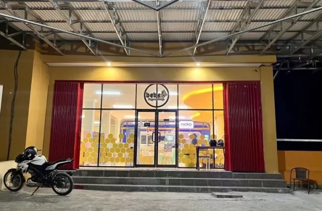 The vibrant yellow facade of Bebe Playground and Cafe in Waingapu is brightly lit at night, with a white motorcycle parked outside its glass entrance.