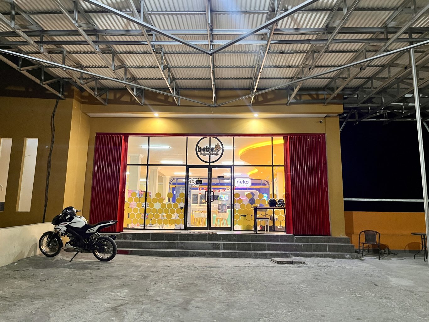 The vibrant yellow facade of Bebe Playground and Cafe in Waingapu is brightly lit at night, with a white motorcycle parked outside its glass entrance.