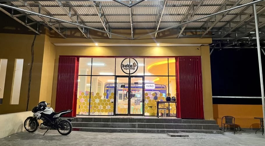 The vibrant yellow facade of Bebe Playground and Cafe in Waingapu is brightly lit at night, with a white motorcycle parked outside its glass entrance.