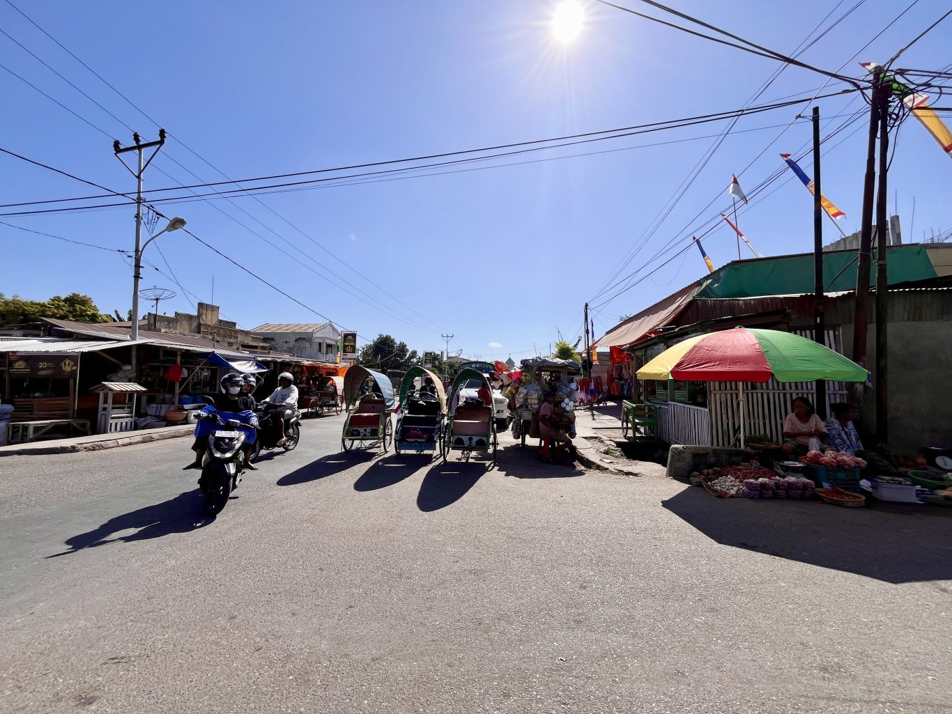 Several traditional becak rickshaws and a scooter are on a sunny street lined with market stalls and buildings under a clear blue sky.