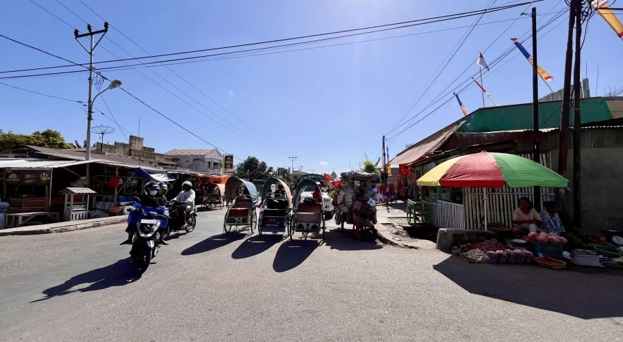 Several traditional becak rickshaws and a scooter are on a sunny street lined with market stalls and buildings under a clear blue sky.