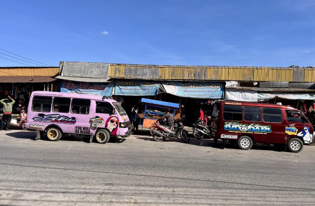Two colorful mini-buses, one pink and one maroon, are parked on a street in front of shops under a clear blue sky, with people and motorcycles nearby.