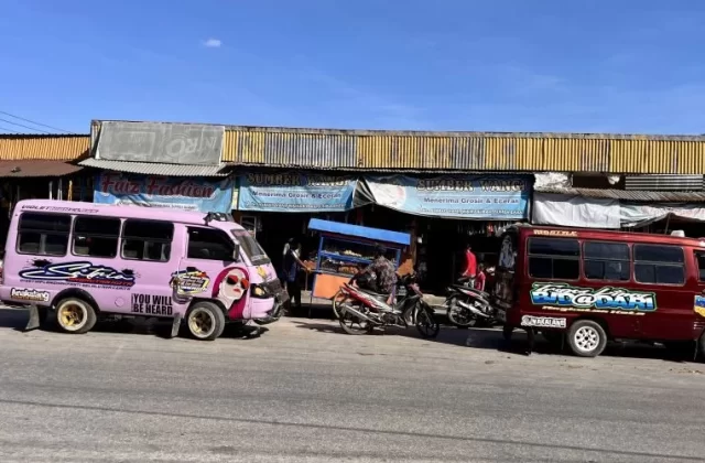 Two colorful mini-buses, one pink and one maroon, are parked on a street in front of shops under a clear blue sky, with people and motorcycles nearby.