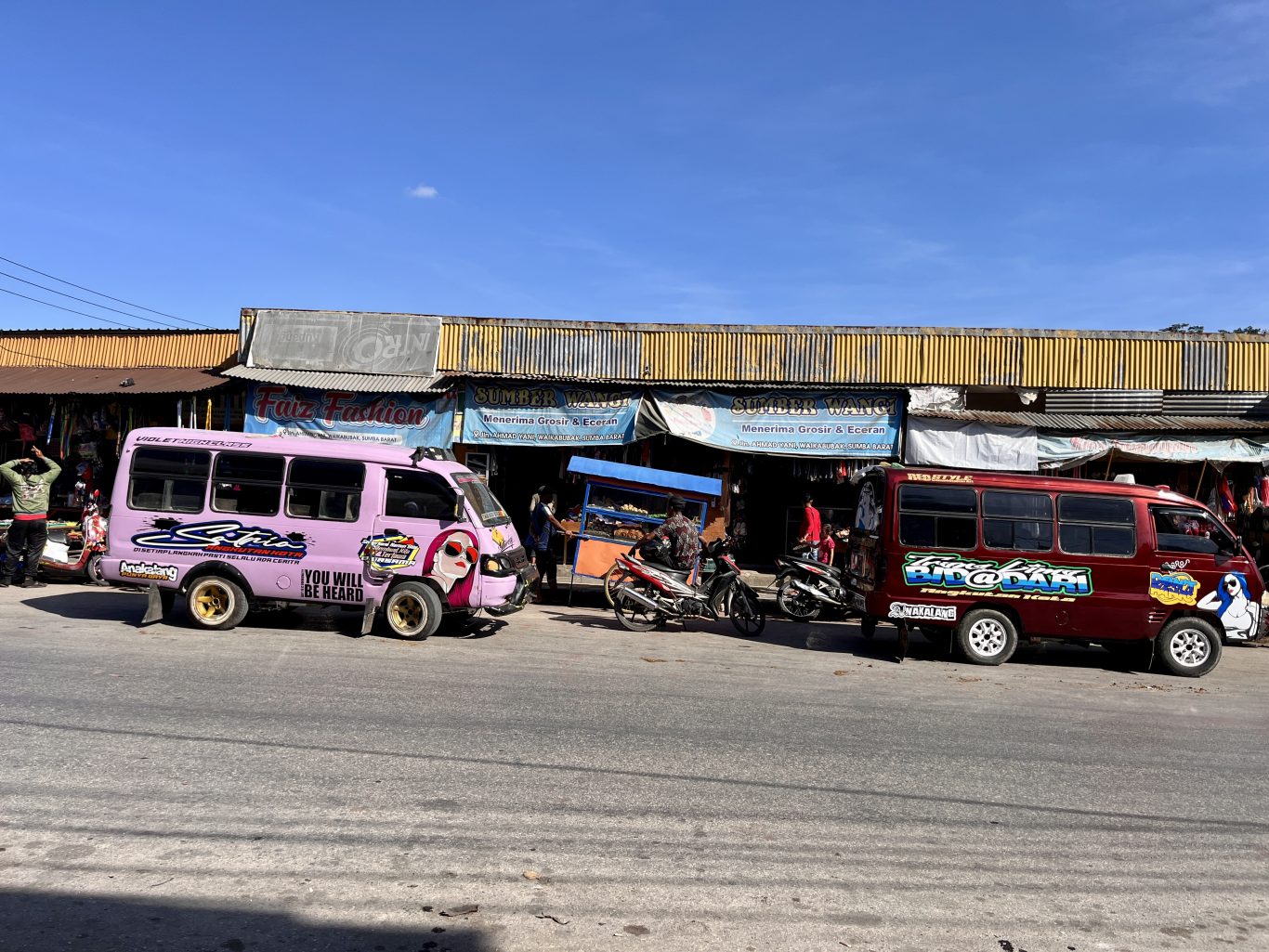 Two colorful mini-buses, one pink and one maroon, are parked on a street in front of shops under a clear blue sky, with people and motorcycles nearby.