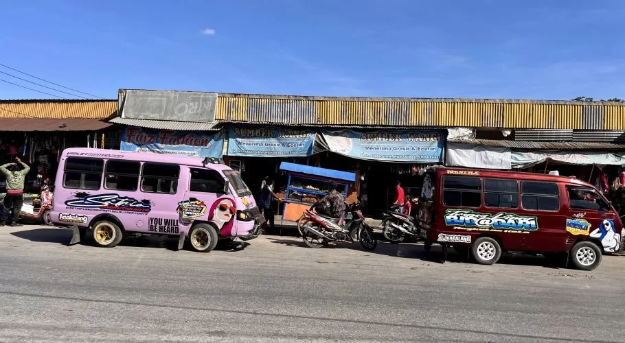 Two colorful mini-buses, one pink and one maroon, are parked on a street in front of shops under a clear blue sky, with people and motorcycles nearby.