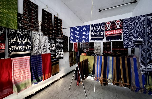 Numerous colorful Sumba ikat textiles with intricate patterns, including horses and geometric designs, hang on white walls and display racks inside a well-lit shop.