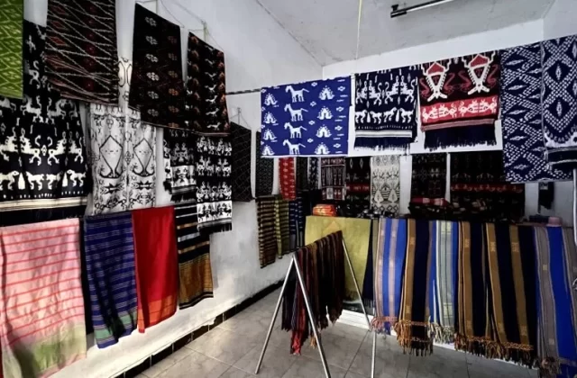 Numerous colorful Sumba ikat textiles with intricate patterns, including horses and geometric designs, hang on white walls and display racks inside a well-lit shop.