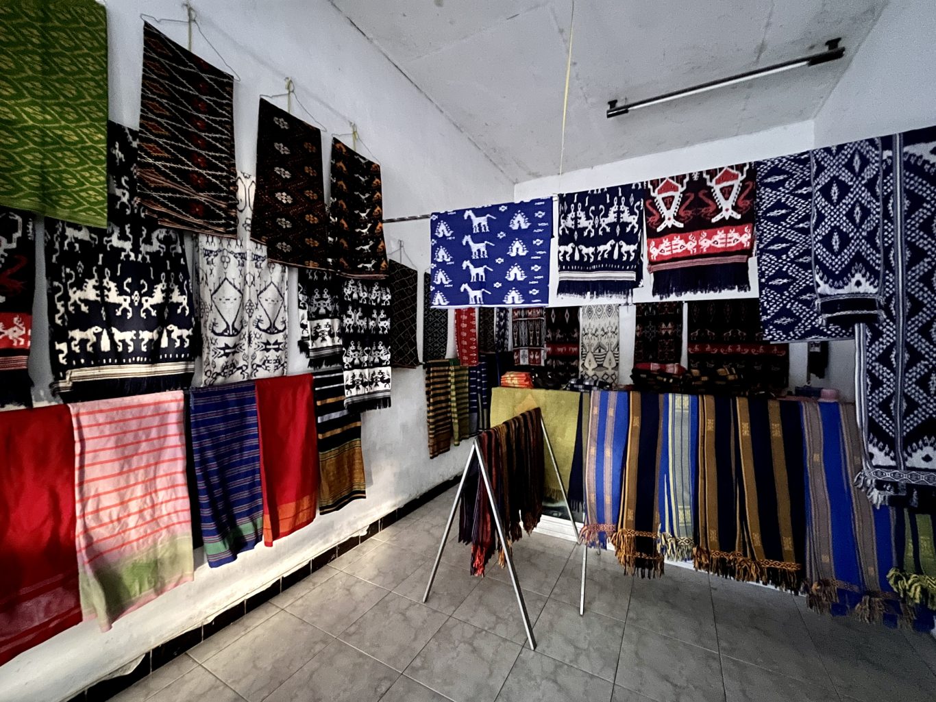 Numerous colorful Sumba ikat textiles with intricate patterns, including horses and geometric designs, hang on white walls and display racks inside a well-lit shop.