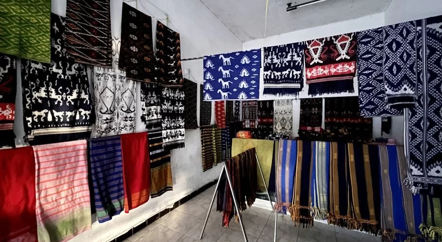 Numerous colorful Sumba ikat textiles with intricate patterns, including horses and geometric designs, hang on white walls and display racks inside a well-lit shop.