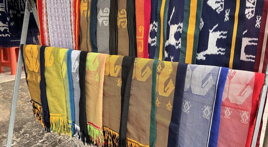 Colorful Sumba ikat textiles with intricate patterns, including deer motifs, hang on display racks in vibrant blues, yellows, reds, and browns.