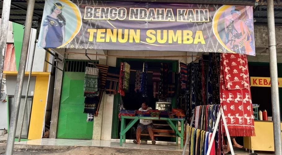 The Bengo Ndaha Kain shop in Tambolaka displays numerous vibrant Sumba ikat textiles, with a man sitting behind a table of folded fabrics.