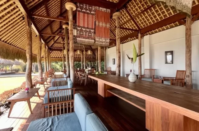 A spacious open-air restaurant features a high thatched roof, many wooden pillars, and a mix of communal and lounge seating with blue cushions.