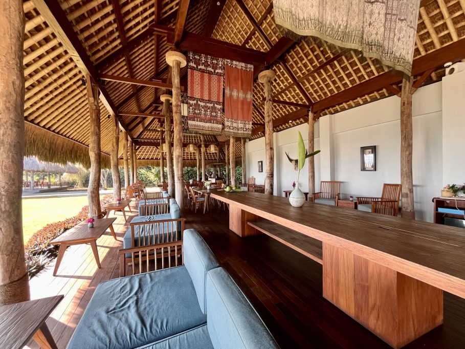 A spacious open-air restaurant features a high thatched roof, many wooden pillars, and a mix of communal and lounge seating with blue cushions.