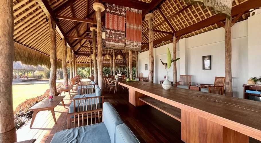 A spacious open-air restaurant features a high thatched roof, many wooden pillars, and a mix of communal and lounge seating with blue cushions.