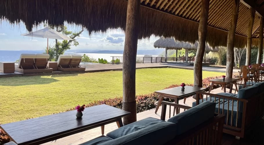 An open-air restaurant with a thatched roof and wooden furniture overlooks a green lawn, sun loungers, and a vast blue ocean under a partly cloudy sky.