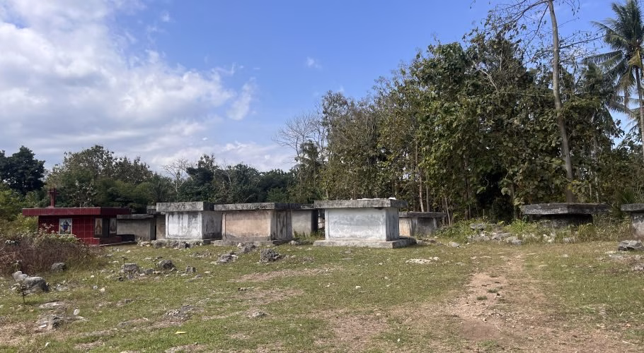 Numerous weathered concrete graves and one red tomb with a cross stand in a grassy field, backed by dense green trees under a blue sky.