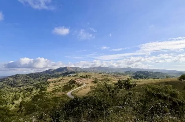 A winding road traverses green and golden savanna hills under a bright blue sky with scattered white clouds, with distant mountains on the horizon.