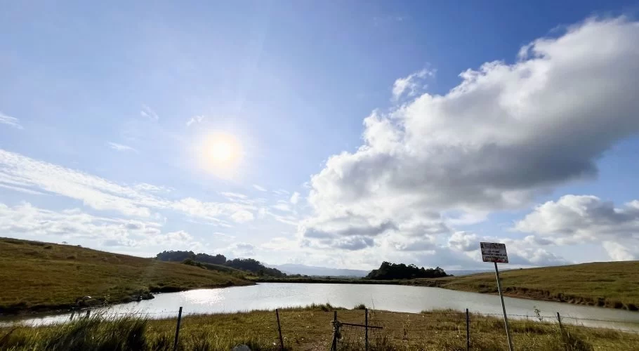 A tranquil lake reflects the bright sun under a blue sky with white clouds, surrounded by rolling green and brown savanna hills.