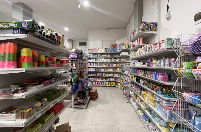 A brightly lit minimarket interior features white shelves filled with groceries, toiletries, and household items, including colorful plasticware and baby products.