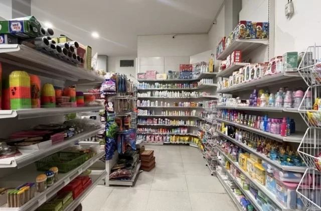 A brightly lit minimarket interior features white shelves filled with groceries, toiletries, and household items, including colorful plasticware and baby products.