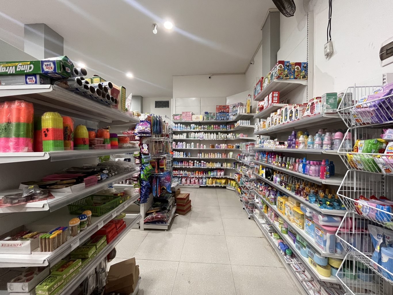 A brightly lit minimarket interior features white shelves filled with groceries, toiletries, and household items, including colorful plasticware and baby products.