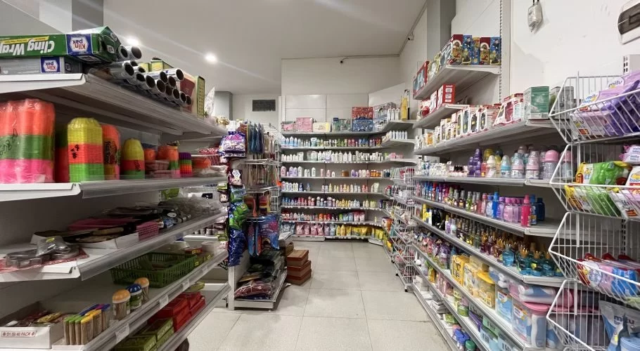 A brightly lit minimarket interior features white shelves filled with groceries, toiletries, and household items, including colorful plasticware and baby products.