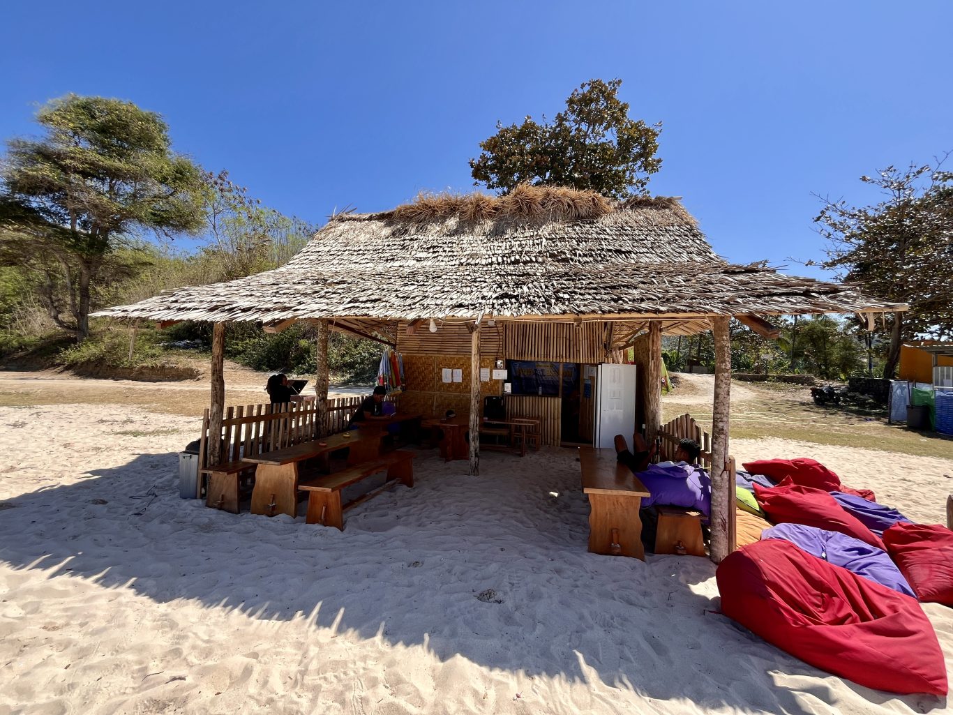 A rustic, open-air restaurant with a thatched roof and wooden tables sits on a sandy beach under a clear blue sky, with trees in the background.