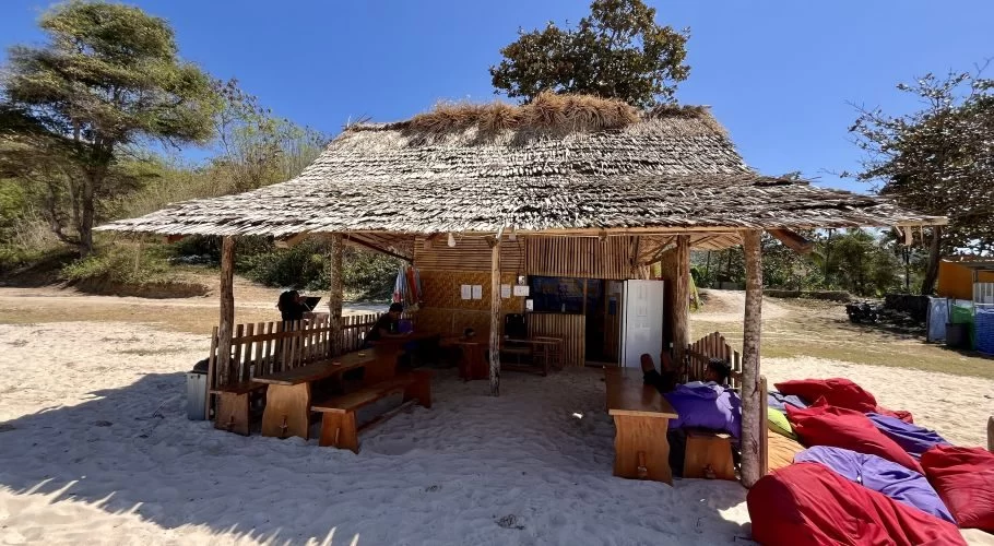 A rustic, open-air restaurant with a thatched roof and wooden tables sits on a sandy beach under a clear blue sky, with trees in the background.