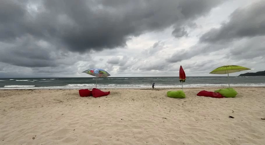 A wide sandy beach under a dark, cloudy sky with three colorful beach umbrellas and beanbags. A person walks near the dark ocean waves.