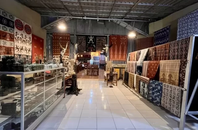 A brightly lit interior of an art shop and cafe features Sumba ikat textiles on walls and racks, with crafts in display cases, leading to a dining area.
