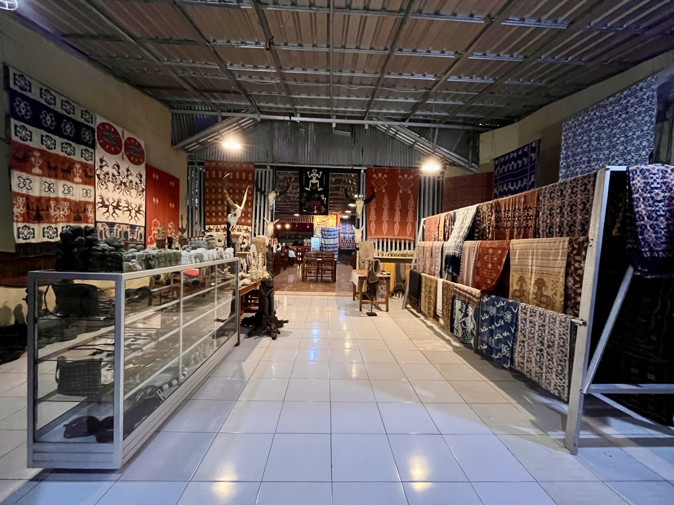 A brightly lit interior of an art shop and cafe features Sumba ikat textiles on walls and racks, with crafts in display cases, leading to a dining area.