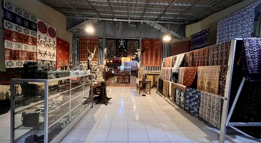 A brightly lit interior of an art shop and cafe features Sumba ikat textiles on walls and racks, with crafts in display cases, leading to a dining area.
