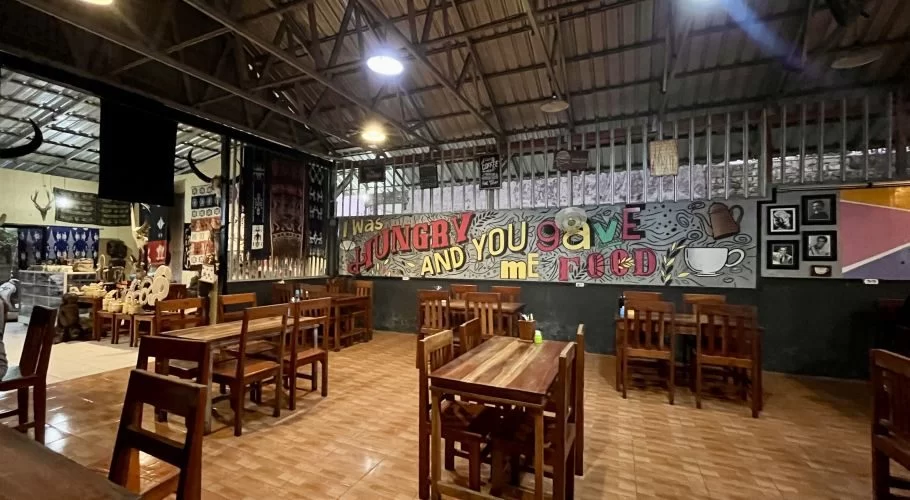 Spacious cafe interior with wooden tables, tiled floor, and a colorful mural. Sumba ikat textiles and artifacts are displayed on the walls.
