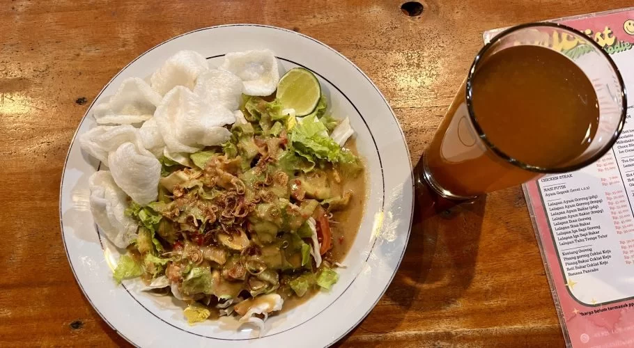 A plate of Indonesian gado-gado with peanut sauce, white crackers, and a lime wedge sits on a wooden table next to a glass of amber tea.