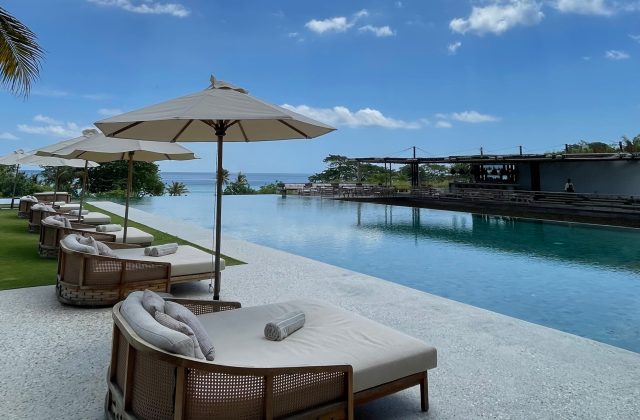 A row of beige sun loungers and umbrellas lines a long infinity pool, with the blue ocean and a resort building visible under a clear sky.