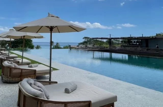 A row of beige sun loungers and umbrellas lines a long infinity pool, with the blue ocean and a resort building visible under a clear sky.