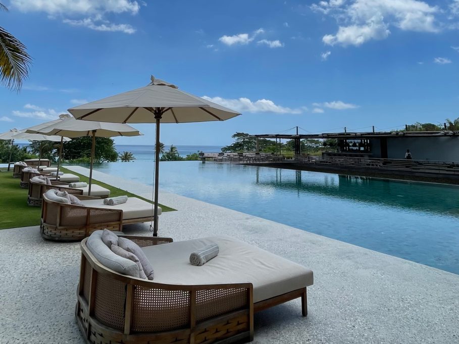 A row of beige sun loungers and umbrellas lines a long infinity pool, with the blue ocean and a resort building visible under a clear sky.