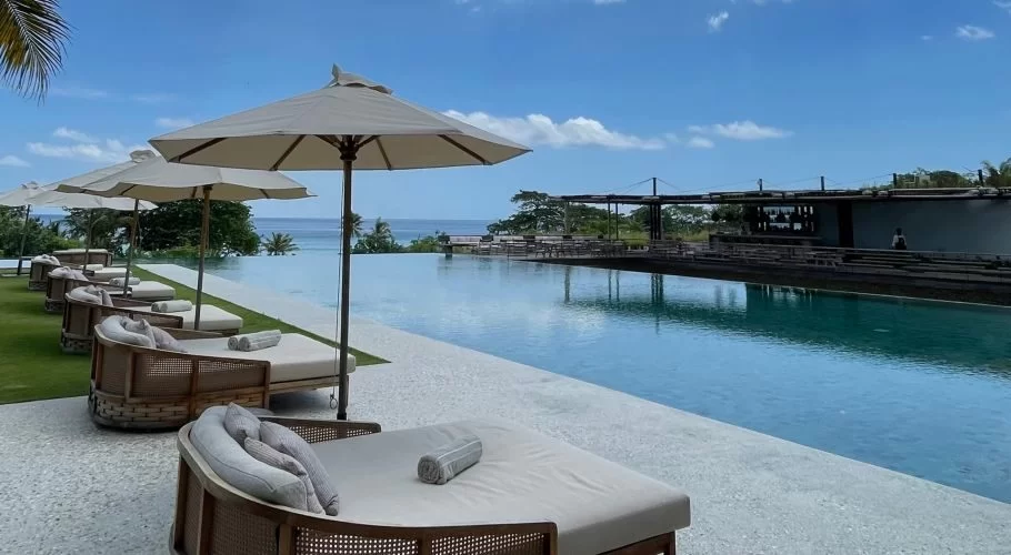 A row of beige sun loungers and umbrellas lines a long infinity pool, with the blue ocean and a resort building visible under a clear sky.