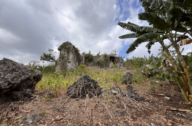 A rugged landscape features large, light-colored rock formations, green foliage, and banana trees under a cloudy sky, with a cave entrance visible.