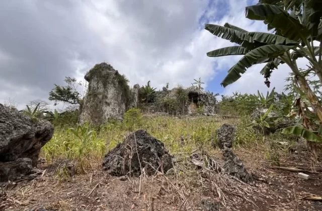 A rugged landscape features large, light-colored rock formations, green foliage, and banana trees under a cloudy sky, with a cave entrance visible.