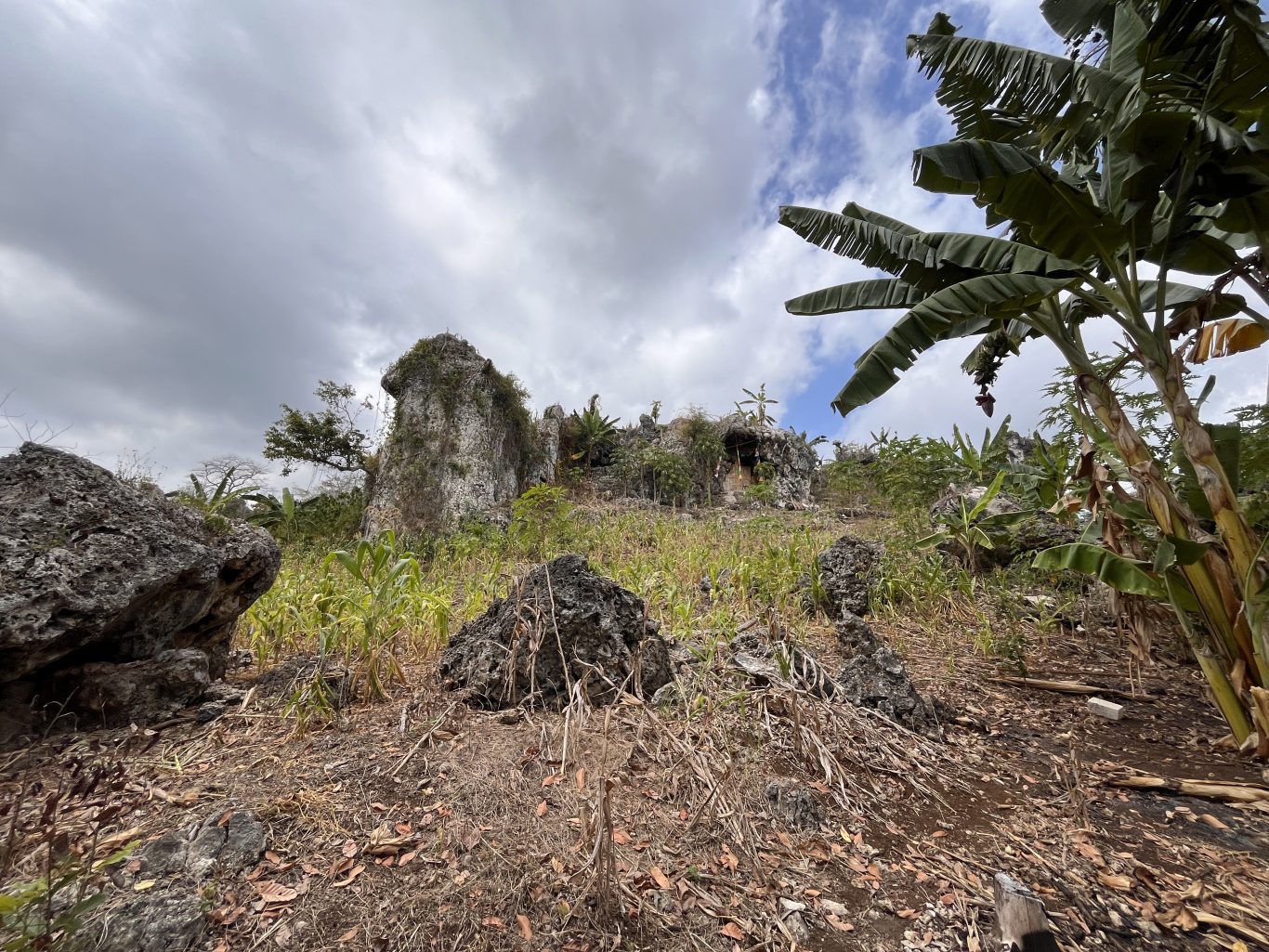 A rugged landscape features large, light-colored rock formations, green foliage, and banana trees under a cloudy sky, with a cave entrance visible.