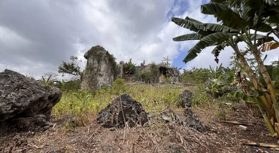 A rugged landscape features large, light-colored rock formations, green foliage, and banana trees under a cloudy sky, with a cave entrance visible.