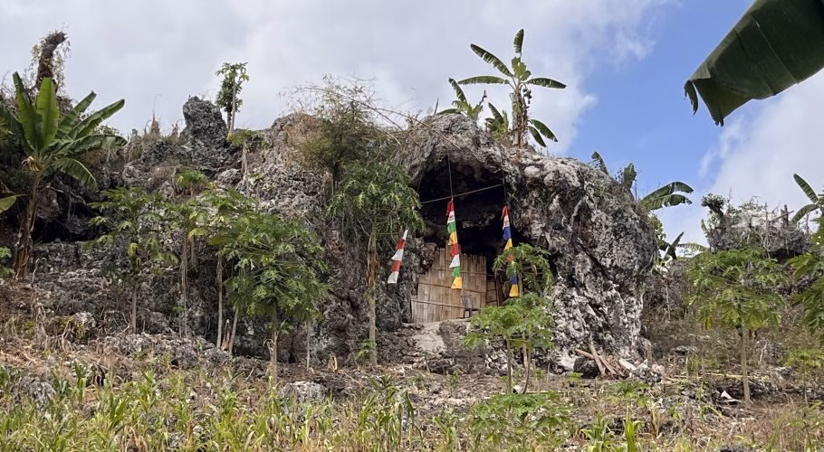 A natural cave entrance with a bamboo door and colorful prayer flags is set into a rocky, green hillside under a partly cloudy sky.