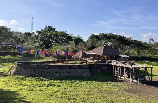 A recreational area in Central Sumba features a colorful 'CEKDAM LOKU UJUNG' sign, thatched huts, and a wooden pier in a green, dry reservoir field under a blue sky.