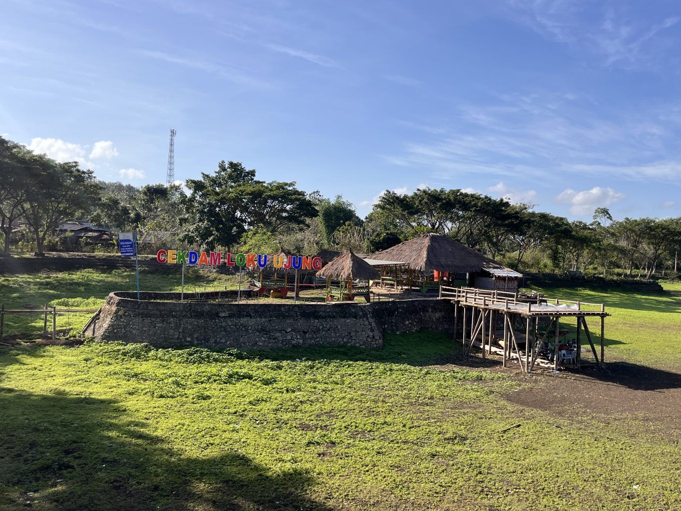 A recreational area in Central Sumba features a colorful 'CEKDAM LOKU UJUNG' sign, thatched huts, and a wooden pier in a green, dry reservoir field under a blue sky.