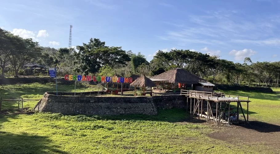 A recreational area in Central Sumba features a colorful 'CEKDAM LOKU UJUNG' sign, thatched huts, and a wooden pier in a green, dry reservoir field under a blue sky.