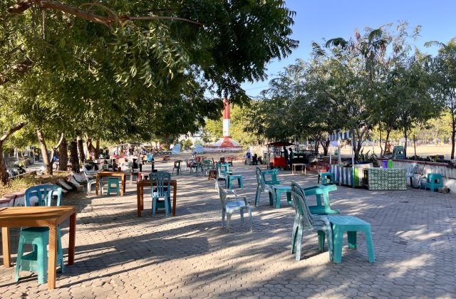 A sunny park scene features many empty plastic tables and chairs under green trees, with vendor stalls and a red and white monument in the background.