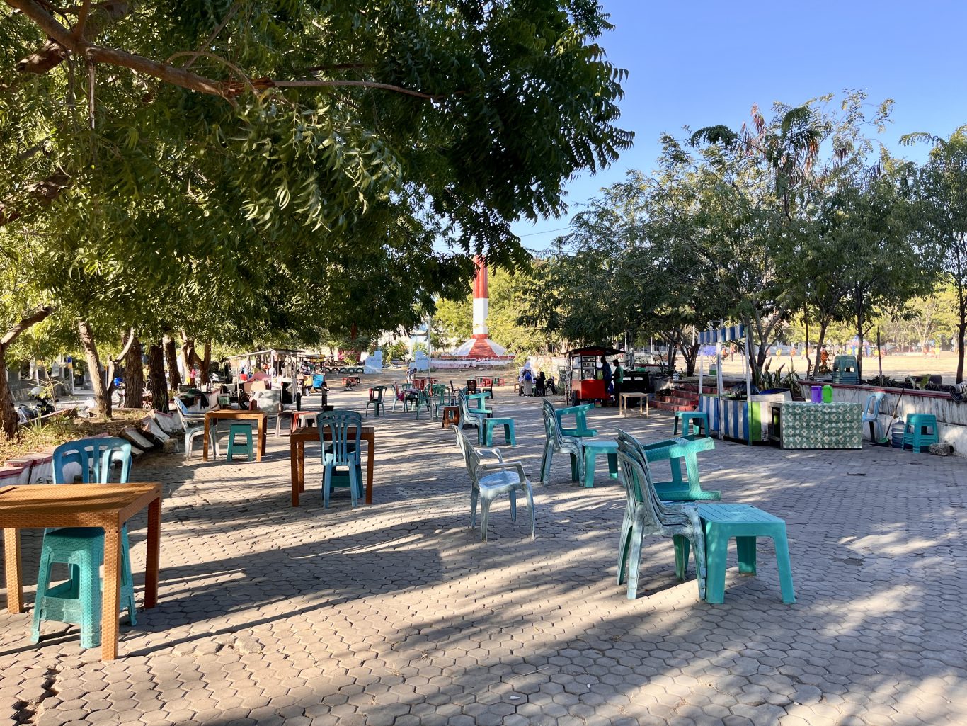 A sunny park scene features many empty plastic tables and chairs under green trees, with vendor stalls and a red and white monument in the background.