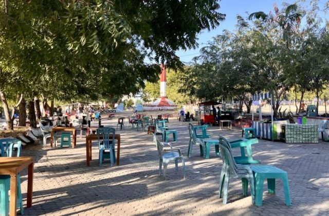 A sunny park scene features many empty plastic tables and chairs under green trees, with vendor stalls and a red and white monument in the background.