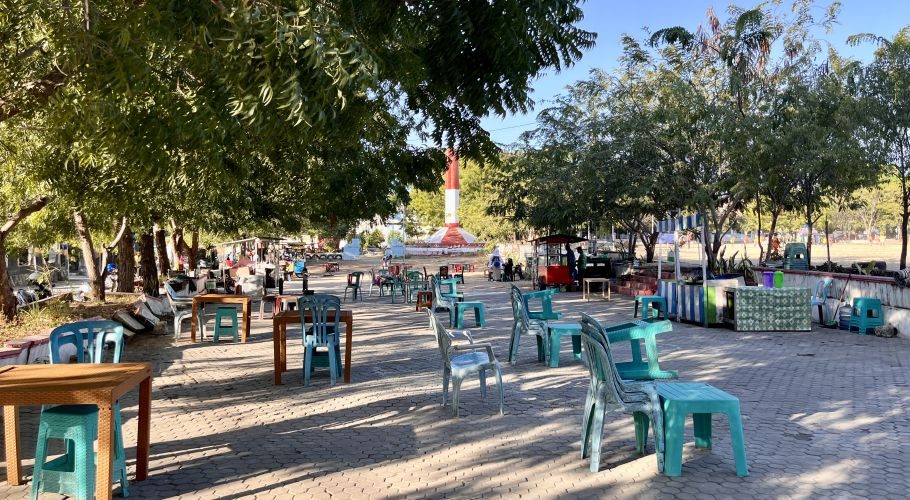 A sunny park scene features many empty plastic tables and chairs under green trees, with vendor stalls and a red and white monument in the background.